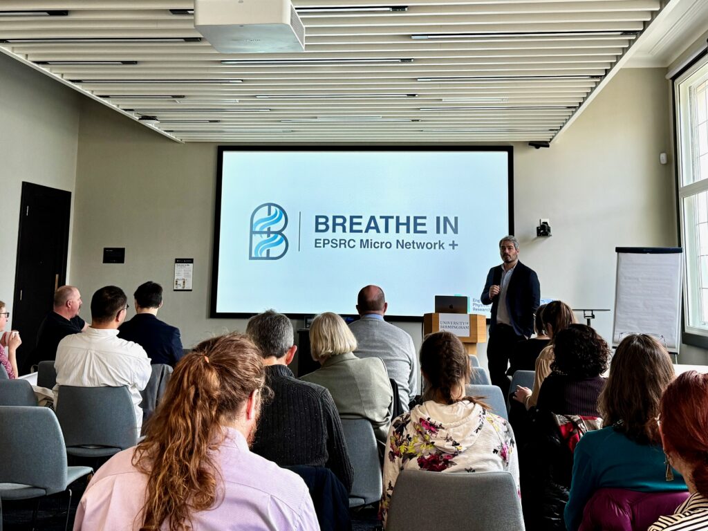 Room where a presentation is being held with a page showing stating 'Breathe in EPSRC micro network+'. Man presenting at front with audience members.